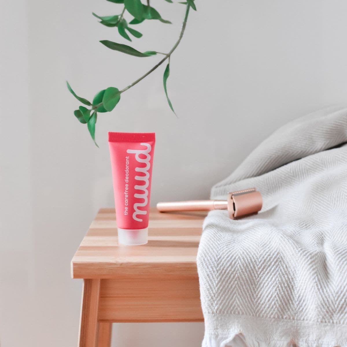 A tube of pink Nuud Deodorant sits on a wooden bedside table next to a gray blanket and a copper-colored roller. Above, a green plant branch adds a touch of nature to the scene, highlighting this natural deodorant that works effortlessly with your lifestyle.