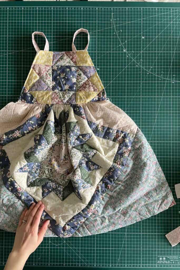 A quilt dress with patchwork rests on a green cutting mat. A hand adjusts the fabric, while a cutting tool peeks in from the right.