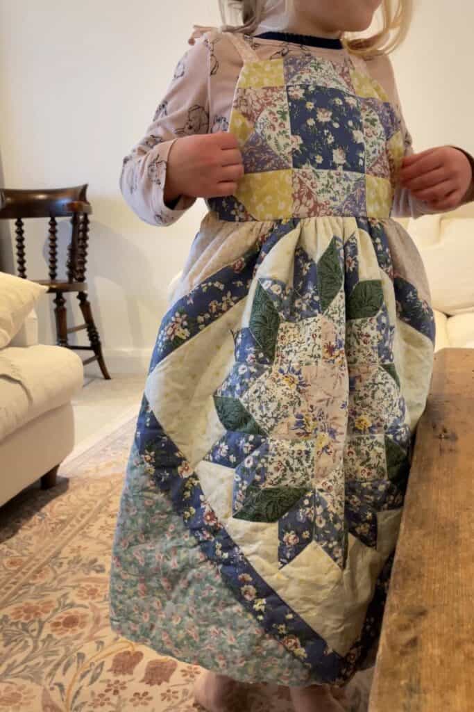 A child wearing a colorful, patchwork dress crafted from an old quilt stands barefoot in a cozy living room. A white couch, a small wooden table, and a patterned rug are visible in the background.