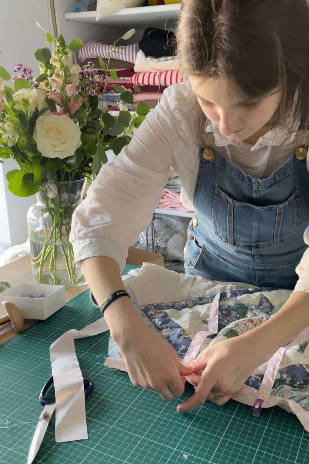 A person in a white blouse and denim overalls is focused on sewing an old quilt on a green cutting mat. Scissors and fabric strips are nearby. A vase with white and pink flowers graces the table, while shelves filled with fabric rolls provide inspiration for a patchwork dress project.