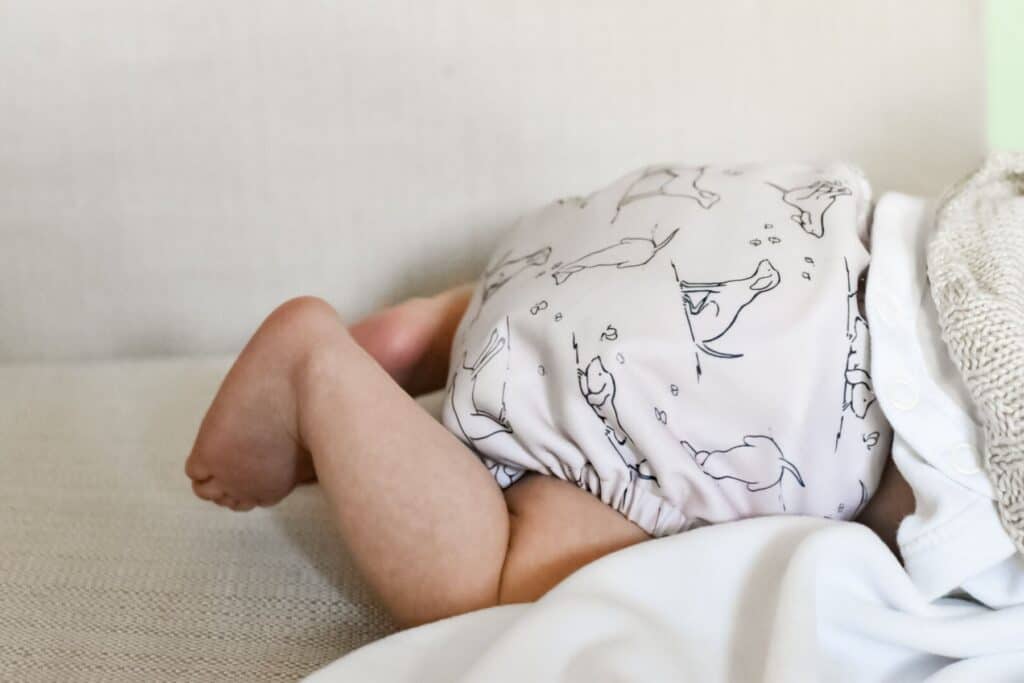 A baby wearing a Peachi Baby Cloth Nappy with animal prints lies on their front on a light-colored sofa. The babys legs are bent, and a white blanket partially covers them.