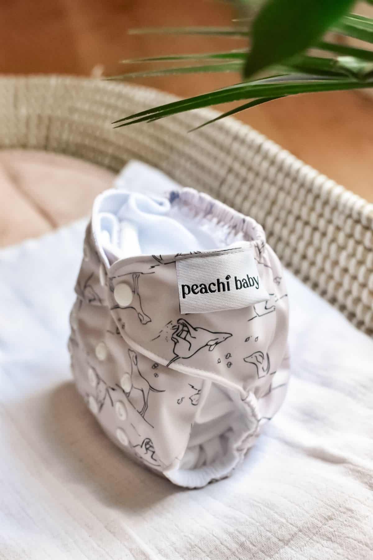 A Peachi Baby cloth nappy rests on a white surface, showcasing a subtle design of abstract shapes and fastened with white snaps. A green leaf peeks from the upper right corner.