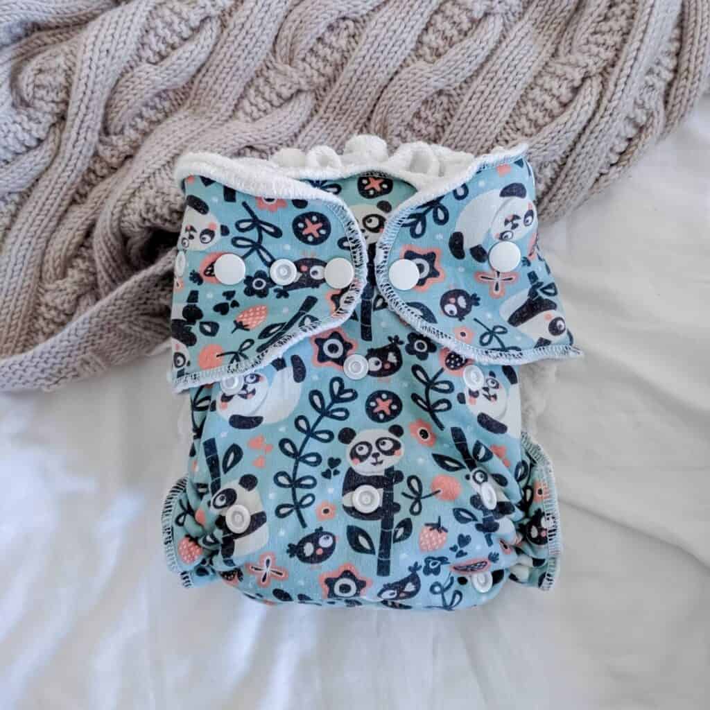 A Petit Lulu Fitted Nappy featuring  a charming panda and floral print in pastel colors, with white snap closures. The cloth nappy sits beside a chunky knit beige blanket.