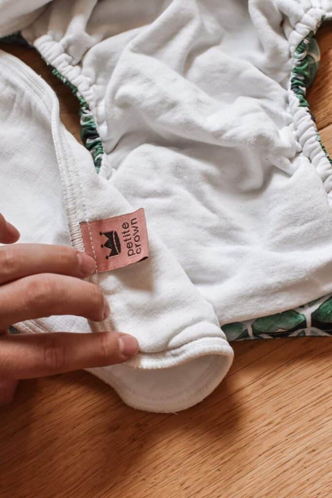 A hand is holding a cloth diaper with a pink tag displaying the Petite Crown logo adorned with a crown. This Trima Nappy, white with green patterned edges, rests on a wooden surface.