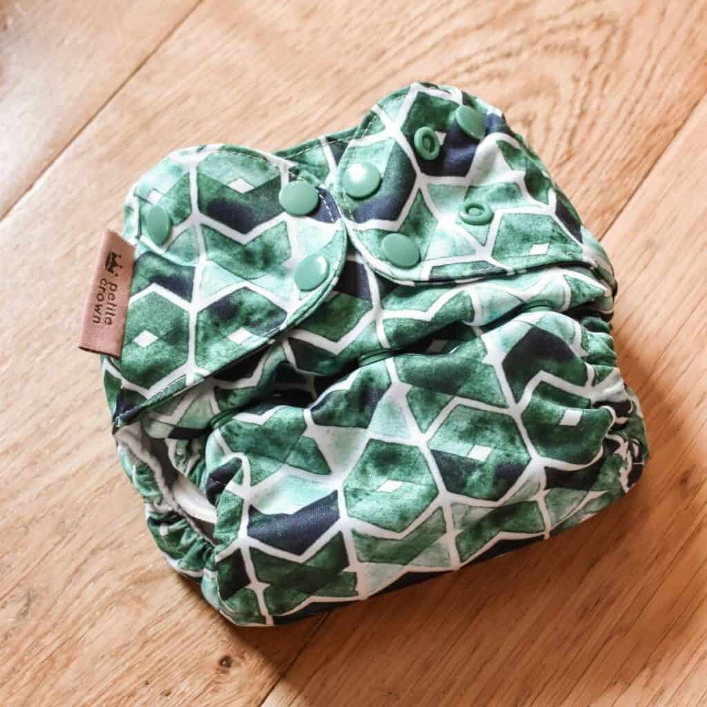 The Petite Crown cloth diaper, known as the Trima Nappy, boasts a green and white geometric pattern with hexagons and diamonds. It features convenient green snap buttons for fastening, lying elegantly on a wooden floor.