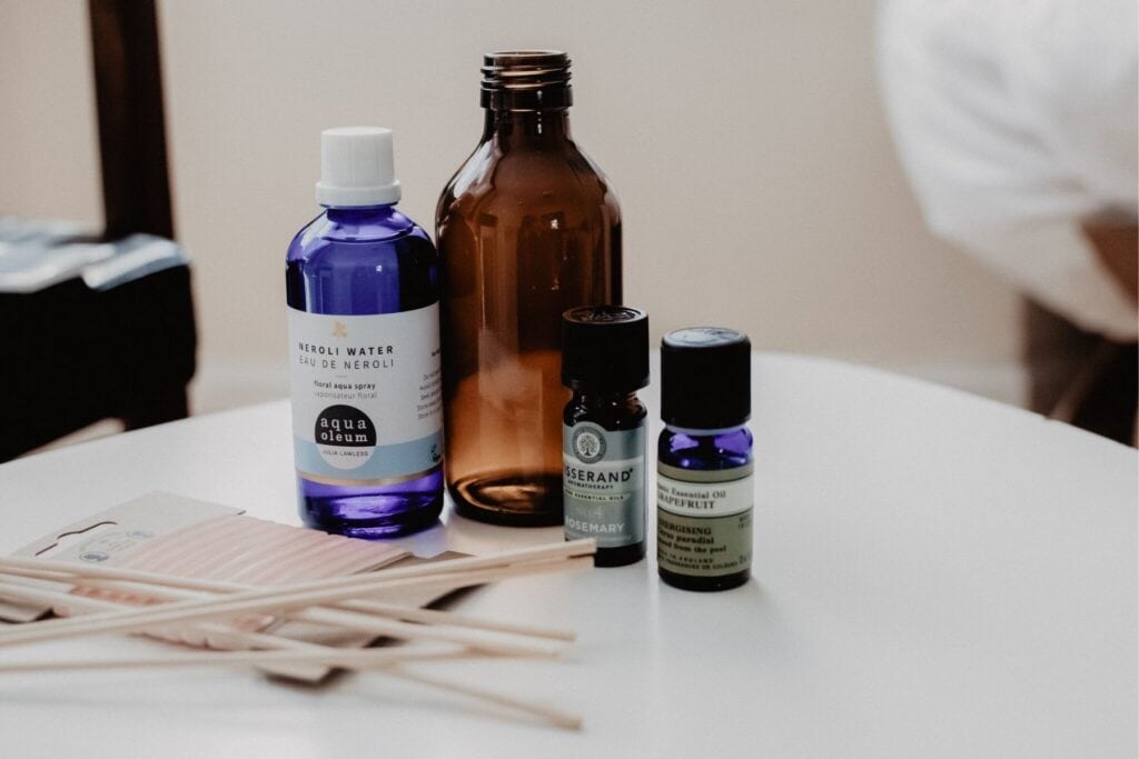A white table showcases various aromatherapy items: a blue bottle of Neroli water, an amber glass bottle, and a small bottle of rosemary essential oil. Diffusing sticks in natural packaging sit elegantly, inviting you to make a high-end reed diffuser. Blurry in the background are a chair and pillow.
