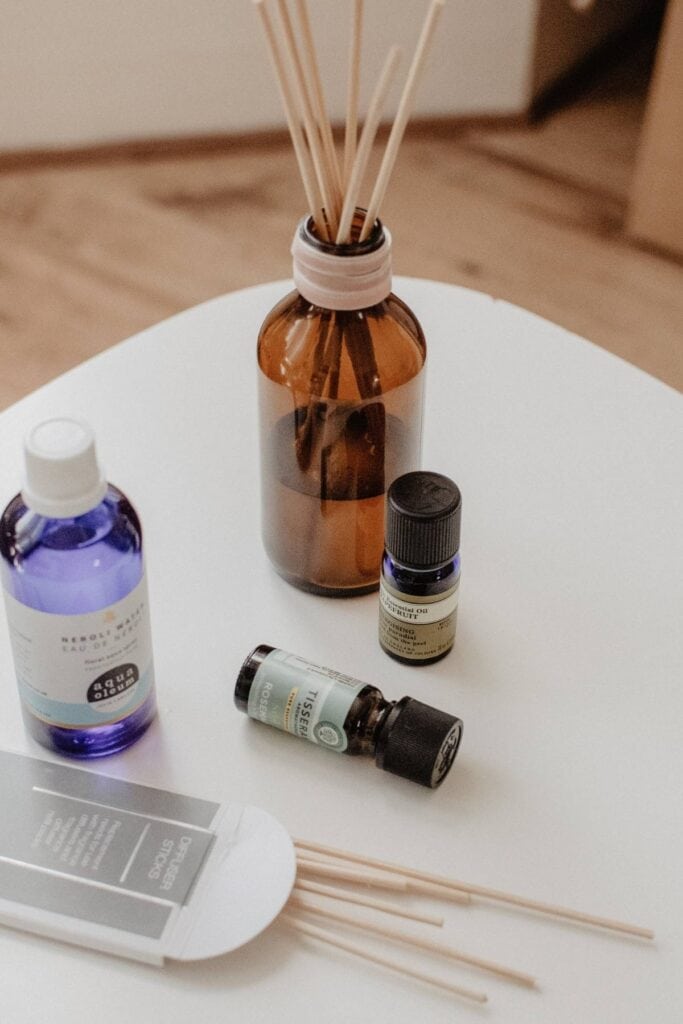 A brown glass reed diffuser bottle with elegant reed sticks stands alongside a blue bottle of essential oil, a sleek black jar, and a black essential oil bottle on a white table. Thin wooden sticks and an inviting package add to the high-end fragrance display.