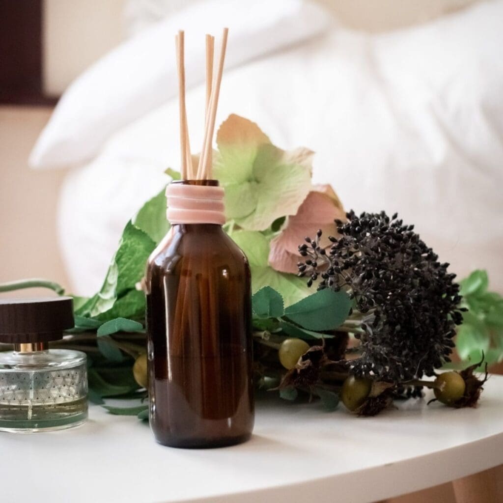 A brown glass reed diffuser bottle with sticks sits on a white table, surrounded by high-end fragrance, artificial green and purple flowers. A small round glass container with a dark lid accompanies it, while a blurred bed softly fades in the background.