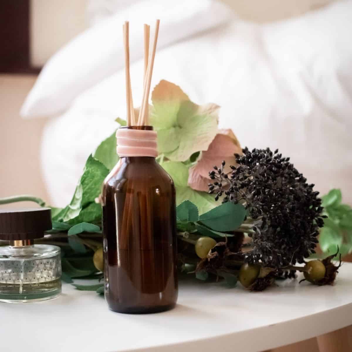 A brown glass reed diffuser bottle with sticks sits on a white table, surrounded by high-end fragrance, artificial green and purple flowers. A small round glass container with a dark lid accompanies it, while a blurred bed softly fades in the background.
