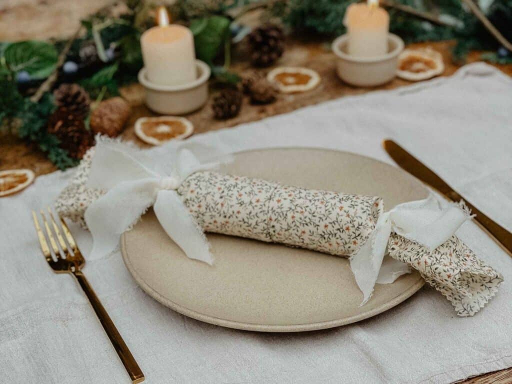 A close-up of a handmade reusable Christmas cracker decorated with a simple white ribbon, resting on a neutral table setting with candles and pinecones in the background.