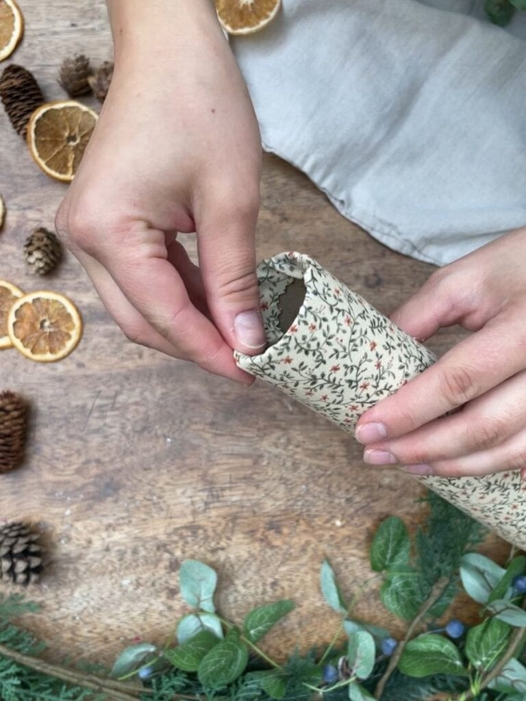 Hands securing the fabric around the cardboard tube, with a cozy, festive atmosphere created by surrounding greenery and dried fruits.