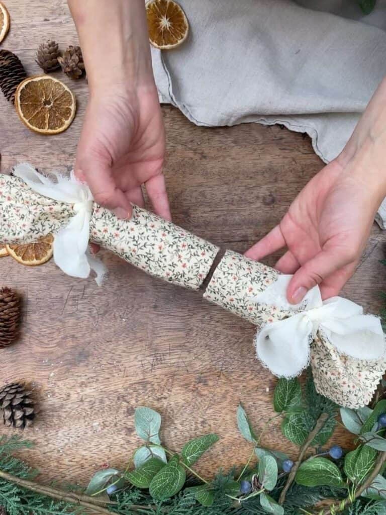 A close-up of a decorated reusable Christmas cracker made from fabric, with a white ribbon tied at both ends, placed on a rustic table surrounded by pinecones, dried orange slices, and greenery.