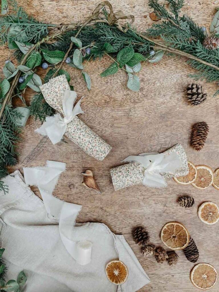 A festive flat lay featuring two reusable Christmas crackers wrapped in floral fabric, surrounded by dried orange slices, pinecones, and greenery.