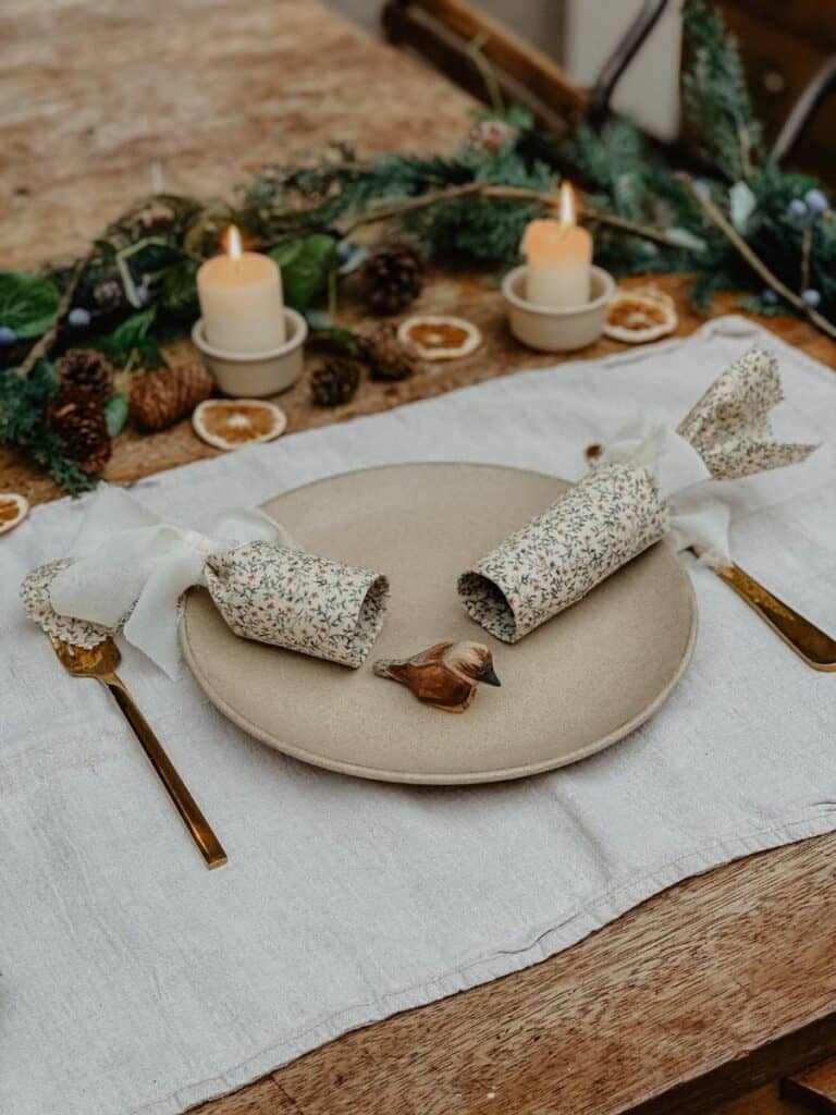 festive Christmas table with a plate and utensils, featuring two handmade reusable Christmas crackers on a linen tablecloth, with pinecones and candles in the background.