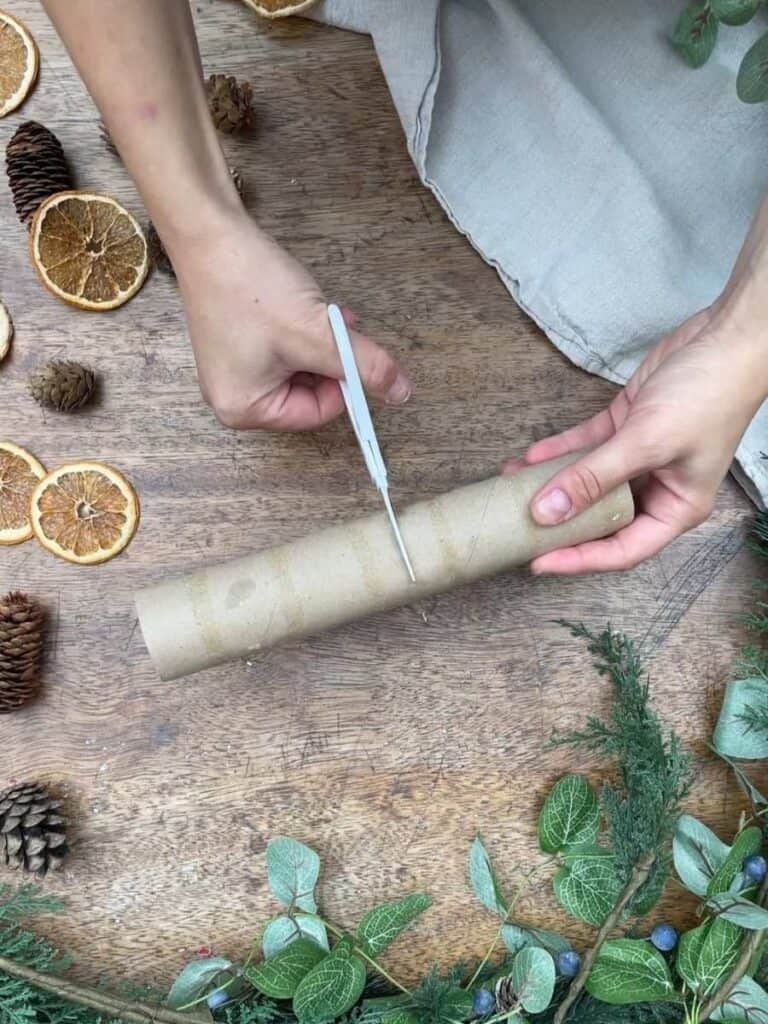 Hands cutting a cardboard tube in the middle, surrounded by festive decorations like dried orange slices and greenery.