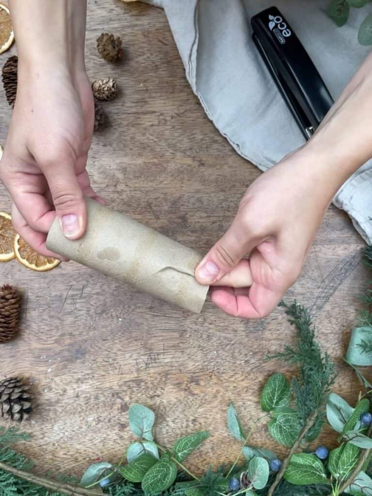 A close-up of hands folding the ends of a cardboard tube, with decorative greenery surrounding the workspace.