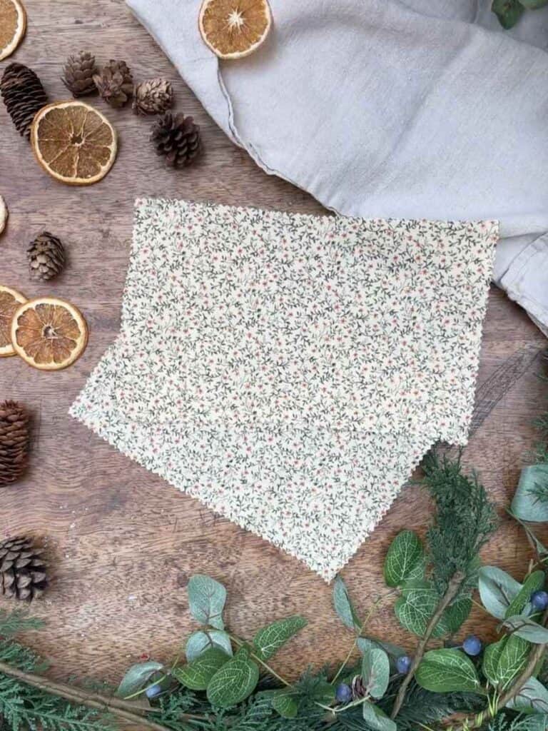 A cut of fabric ready to be used to make reusable Christmas crackers, surrounded by festive décor such as pinecones, dried oranges, and greenery on a wooden table.