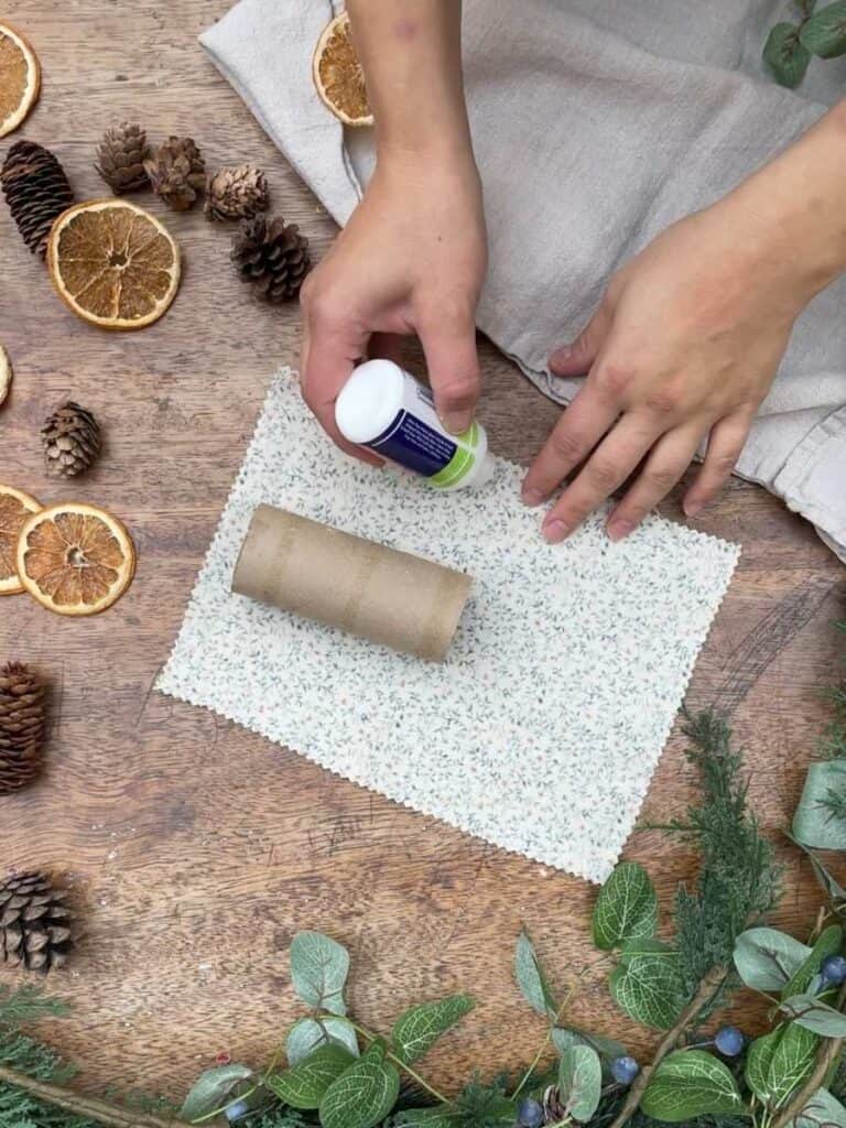A pair of hands gluing floral fabric along one edge, with festive decorations like pinecones and dried oranges in the background.