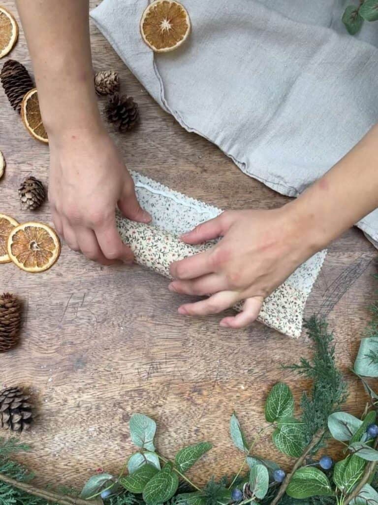 Hands carefully rolling floral fabric around a cardboard tube, beginning the creation of a reusable Christmas cracker, with dried oranges and pinecones on the table.