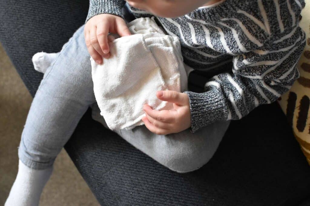 A child in a striped sweater and gray pants sits on a dark couch with legs crossed, carefully holding a stack of reusable wipes. Only the upper body and hands are visible.