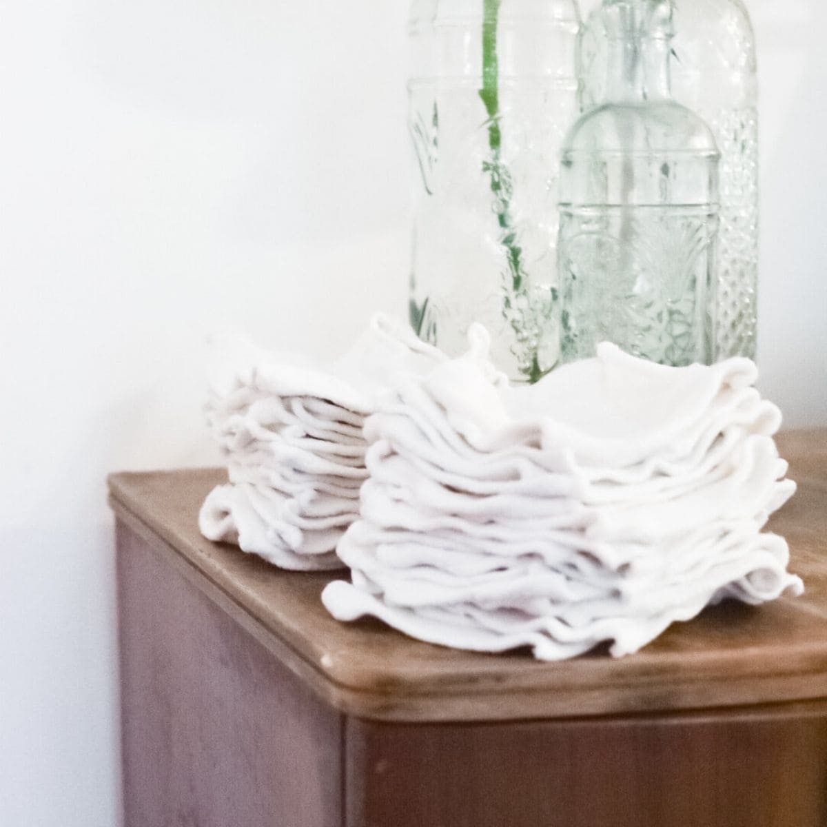 A stack of neatly folded reusable wipes is placed on a wooden surface. Behind them are three clear glass bottles with etched designs, reflecting the light softly against a plain white background.