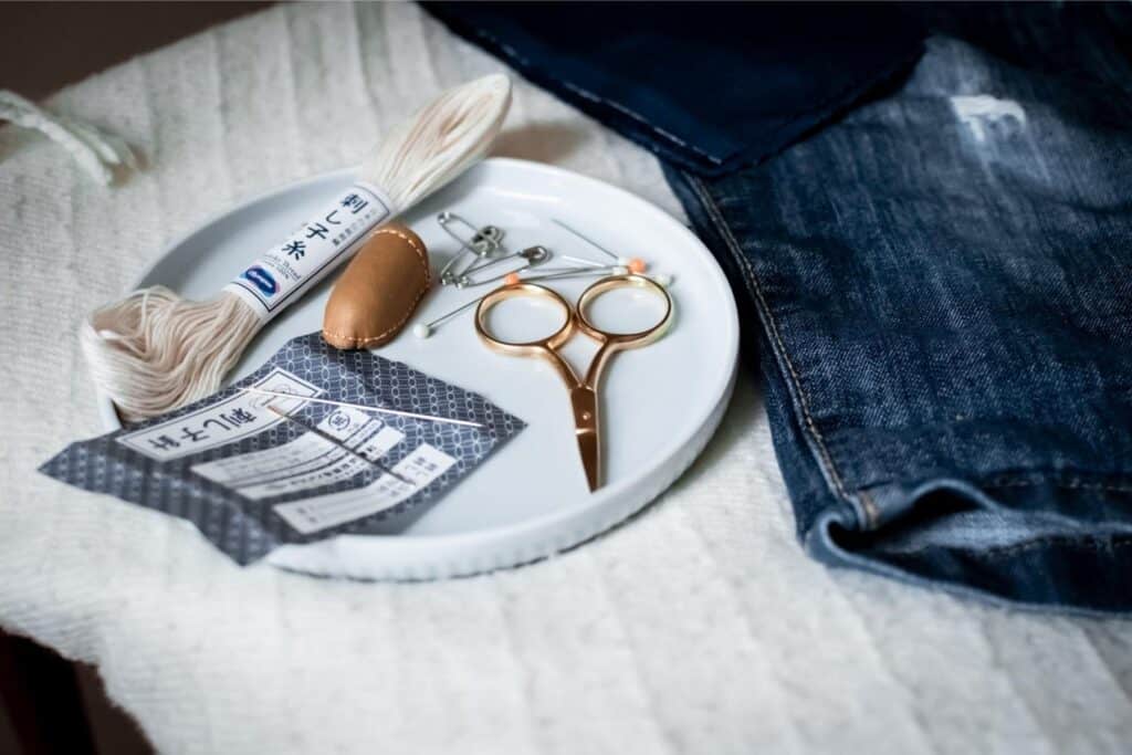 A white plate holds sewing tools, including a pair of scissors, safety pins, sashiko thread, and a thimble. Nearby, denim fabric pieces are ready to be mended.