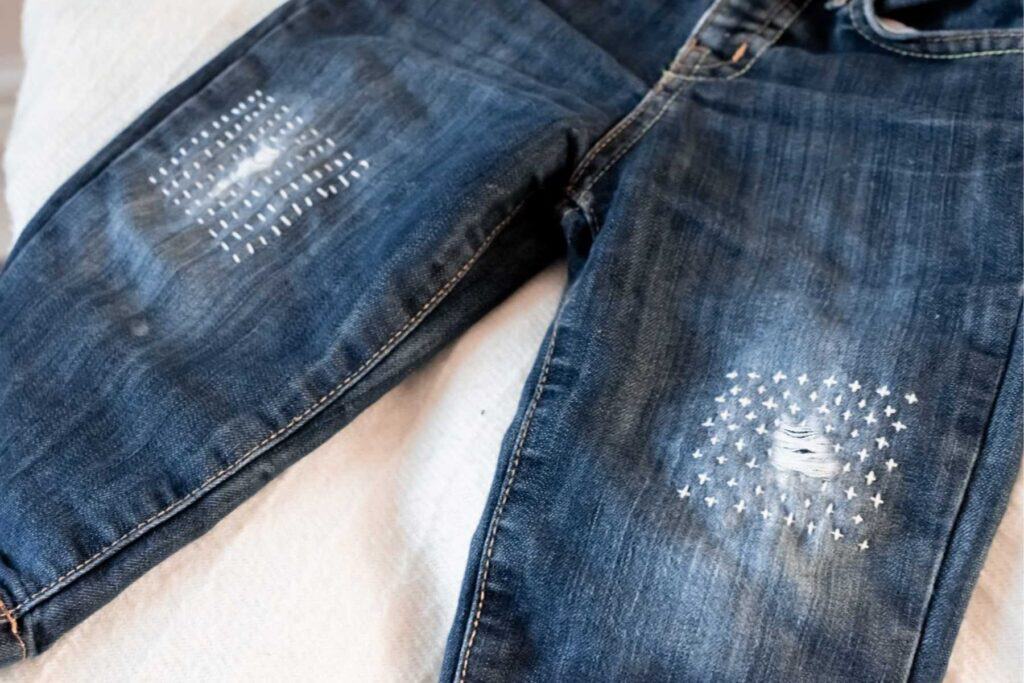 Close-up of a pair of dark blue jeans with visible white Japanese sashiko stitching repairs on both knees, forming cross patterns. The jeans are laid flat on a light-colored surface.