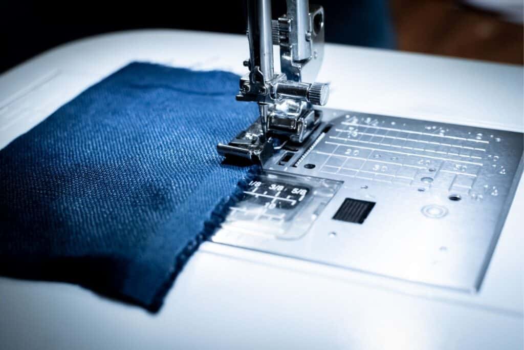Close-up of a sewing machine in action, overlocking the edge of a patch of blue fabric. The needle and presser foot are visible, along with the metal plate marked with measurement lines. 