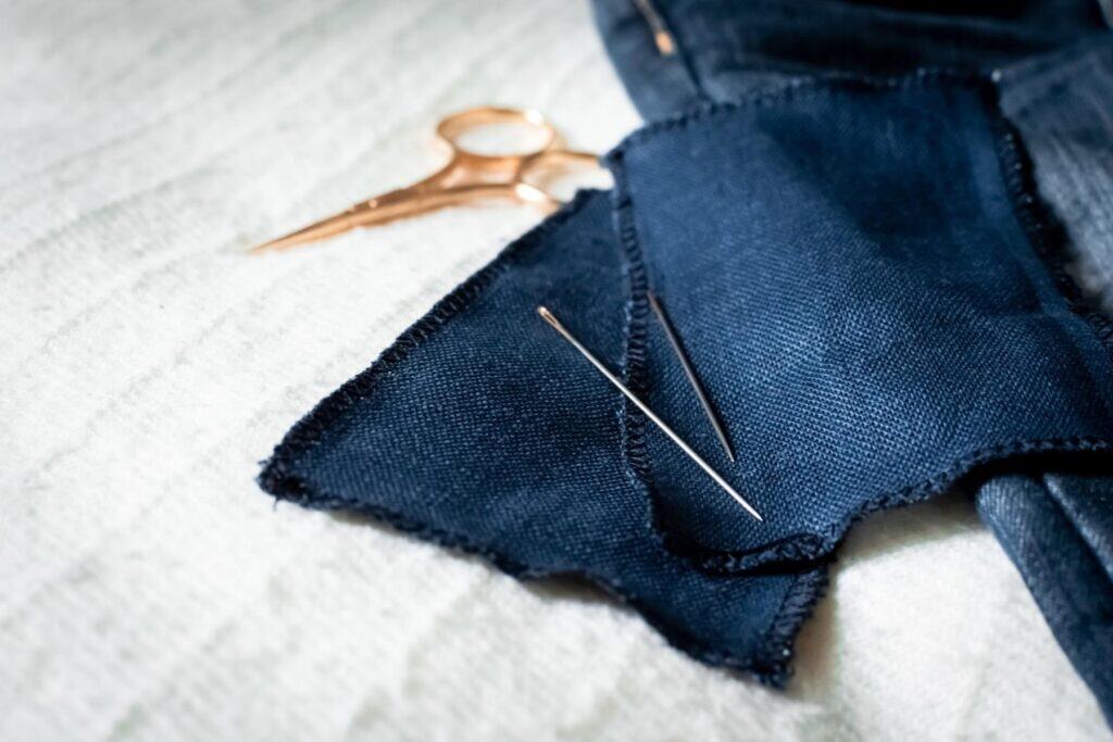Close-up of a dark blue fabric patch with two sashiko needle poised on top, beside gold-colored scissors on a light-colored surface.
