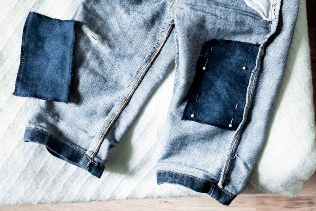 A pair of blue jeans, inside out, with dark blue Sashiko patches pinned onto the legs lies on a cream-colored surface. 