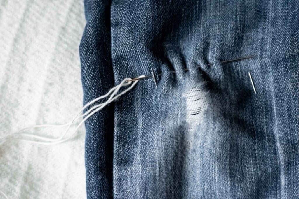 A close-up of blue denim jeans with a small tear being mended using Sashiko, the Japanese stitching technique. A needle with white thread weaves in and out around the tear, demonstrating the artful process of hand-stitching on a textured, light-colored surface.