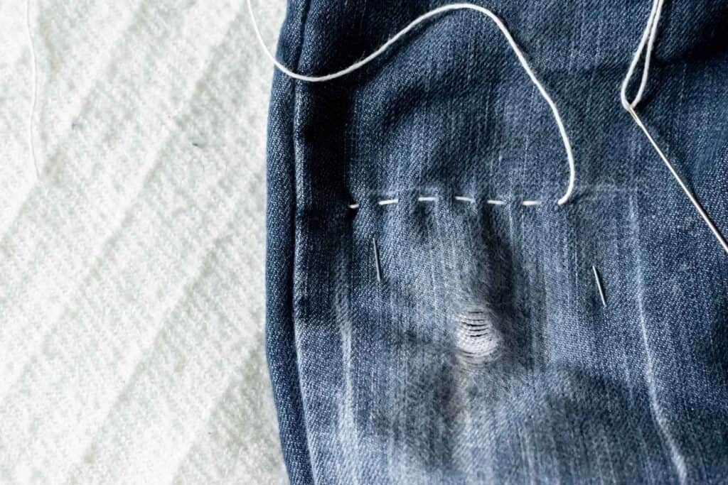 Close-up of denim fabric showcasing the Sashiko technique with visible hand-stitching and a threaded needle. The parallel stitches run in a straight line above the small hole. 