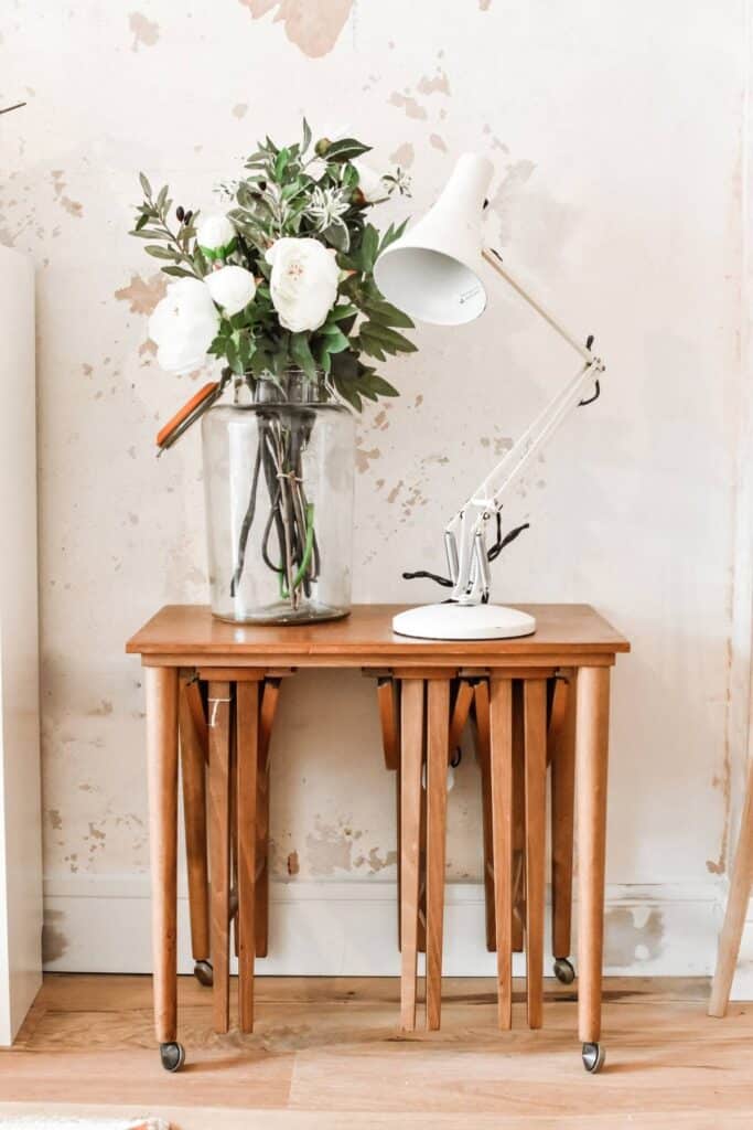 A secondhand wooden nesting table holds a bouquet of white flowers in a clear vase and a white adjustable lamp. The table is set against a textured, light-colored wall. The wooden floor enhances the rustic ambiance.