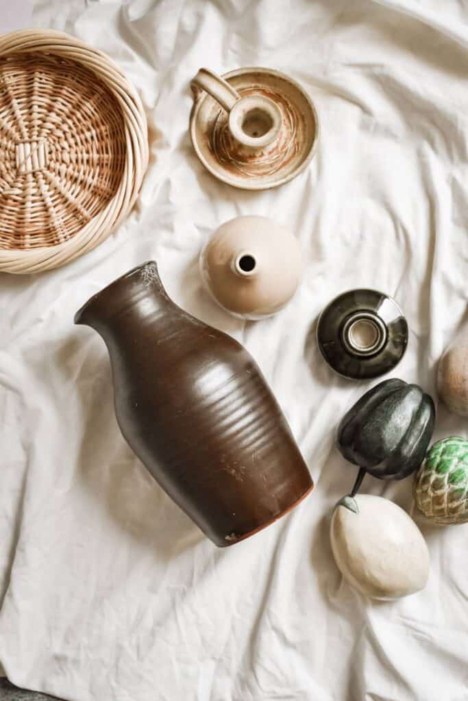 A curated collection of secondhand ceramic items on a white fabric surface, featuring a large brown vase, a small tan teapot, and a round black vase, alongside a basket and several decorative gourds and vessels in earthy tones.