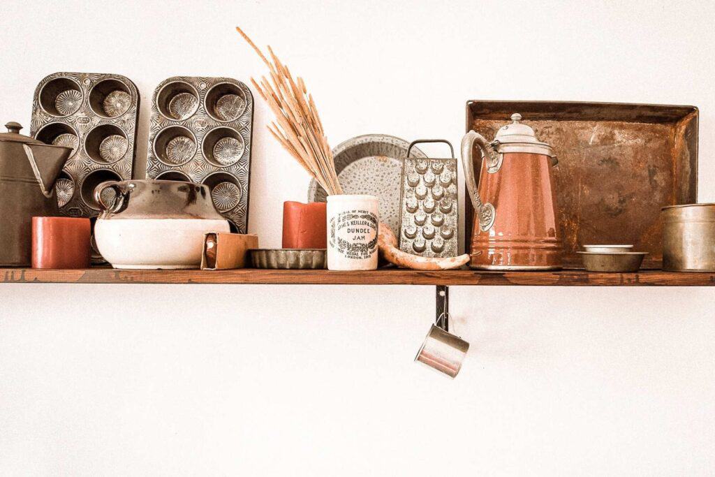 A wooden shelf showcases vintage kitchenware treasures from secondhand shopping, featuring muffin tins, a teapot, baking trays, and a mix of ceramic and metal containers. A small potted plant and a hanging cup add charm to this unique collection.
