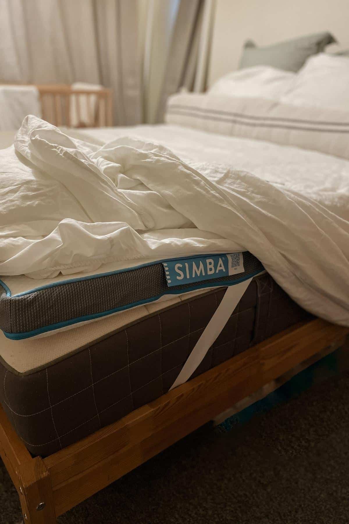 A cozy bedroom scene with an unmade bed featuring a white comforter and pillows. The bed has a visible SIMBA mattress topper on a wooden frame. In the background, another bed or crib is partially visible, and soft curtains hang by the window.
