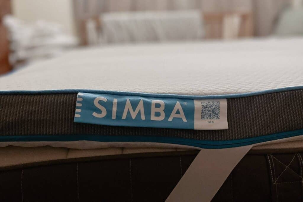 Close-up of a mattress topper with a blue and white label on the side that reads SIMBA. A QR code is visible next to the text. The background shows a blurred view of furniture and stacked linens.