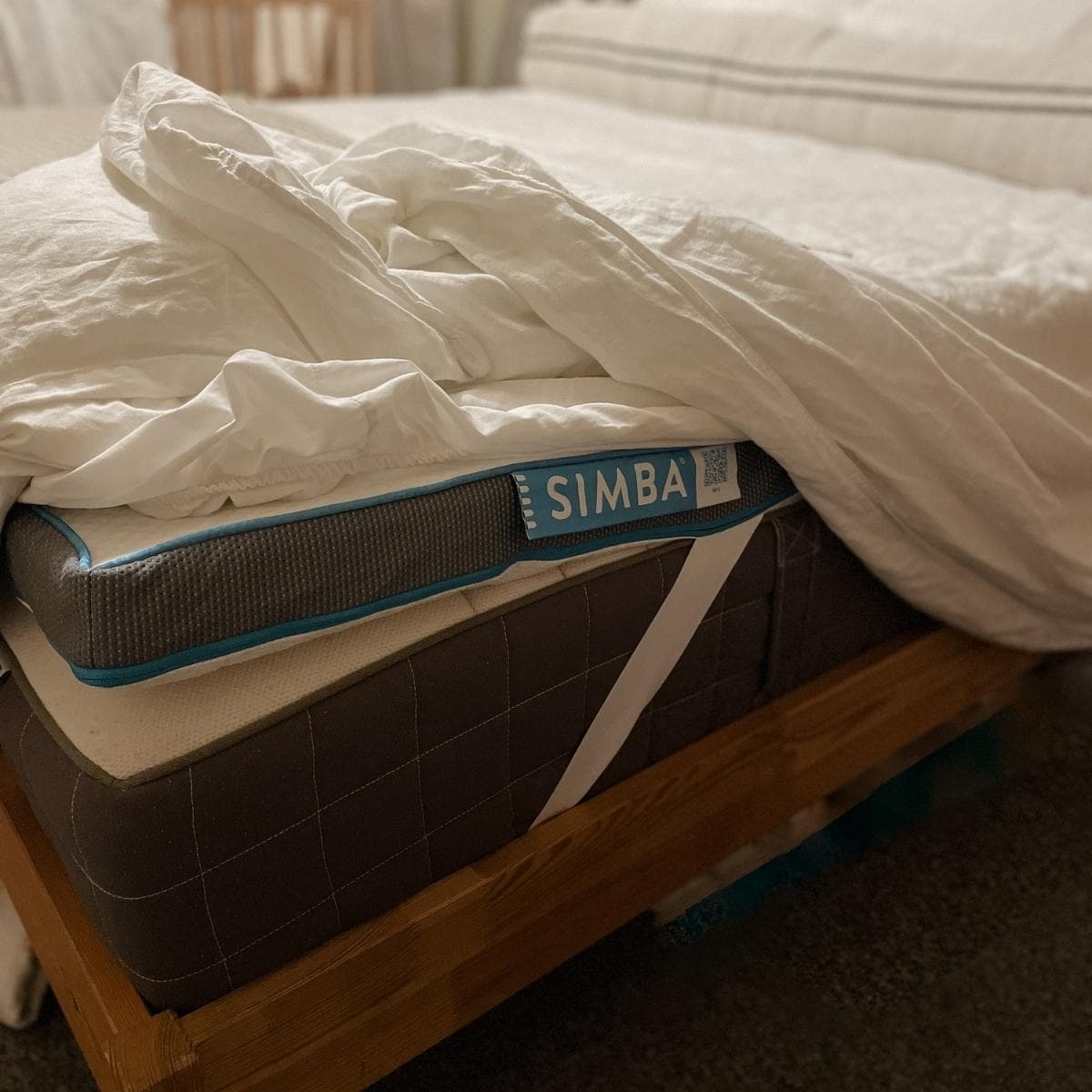 A bed with a neatly tucked white comforter and a visible mattress topper. The mattress topper has a blue edge and SIMBA branding. The bed frame is wooden, and the floor is carpeted.