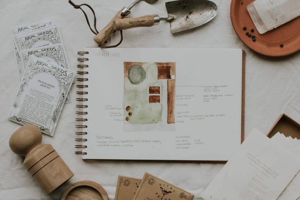 A sketchbook open to a 2023 small cottage garden plan with a watercolor layout is surrounded by gardening tools, seed packets, and a wooden tamper. A trowel and string are nearby.