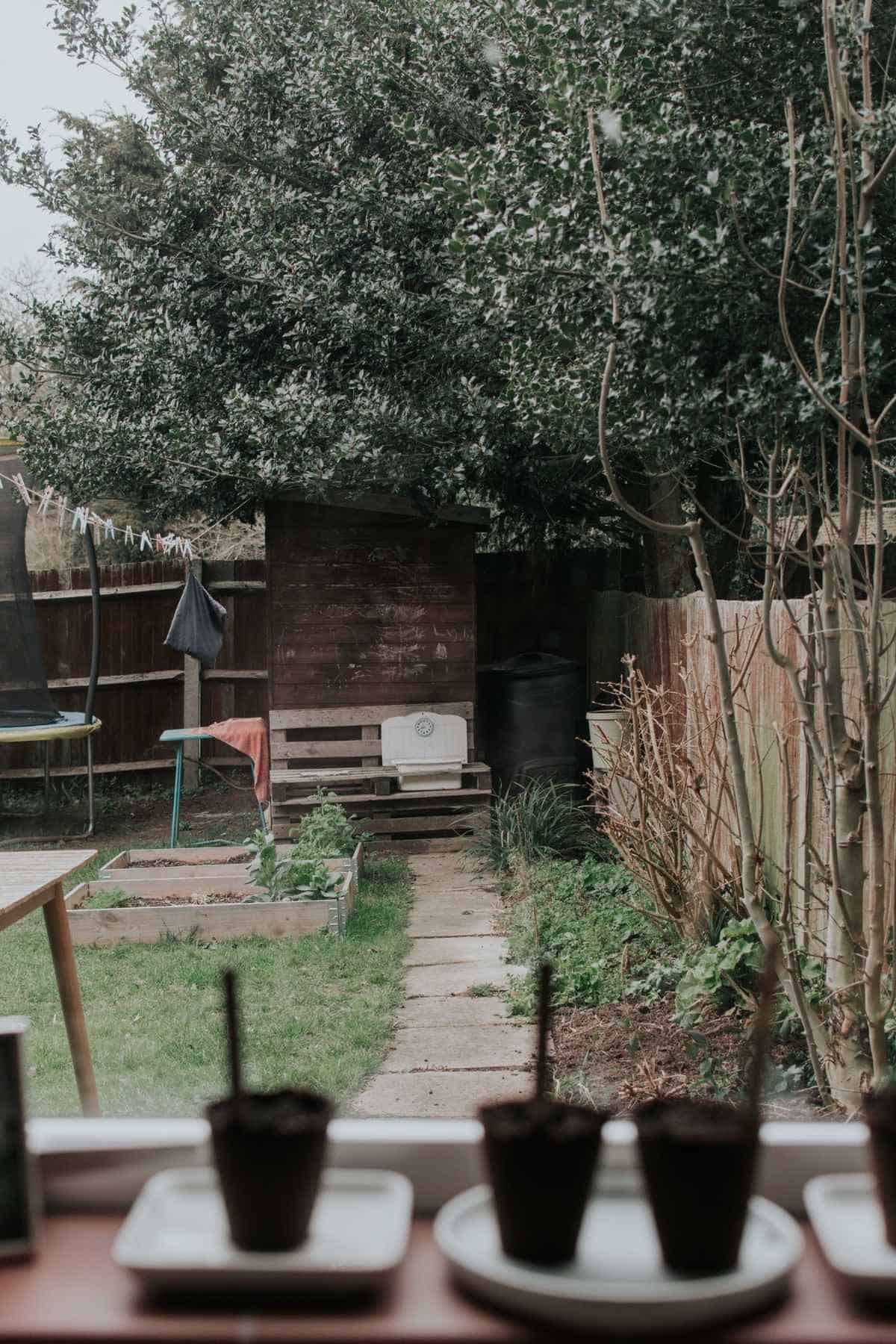 A view from the window reveals a small cottage garden with a stone path leading to a wooden shed. Leftward, a trampoline sits beside two raised beds. Three small potted plants rest on plates along the window ledge, perfect plans for cultivating charm in compact spaces.