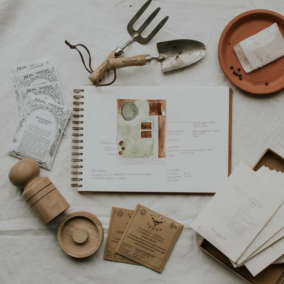 A flat lay of garden planning items on a white cloth reveals plans for a small cottage garden. Includes a sketchbook with a 2023 design, seed packets, a trowel, a gardening fork, and wooden containers. Theres also a small plate with seeds and a piece of paper.