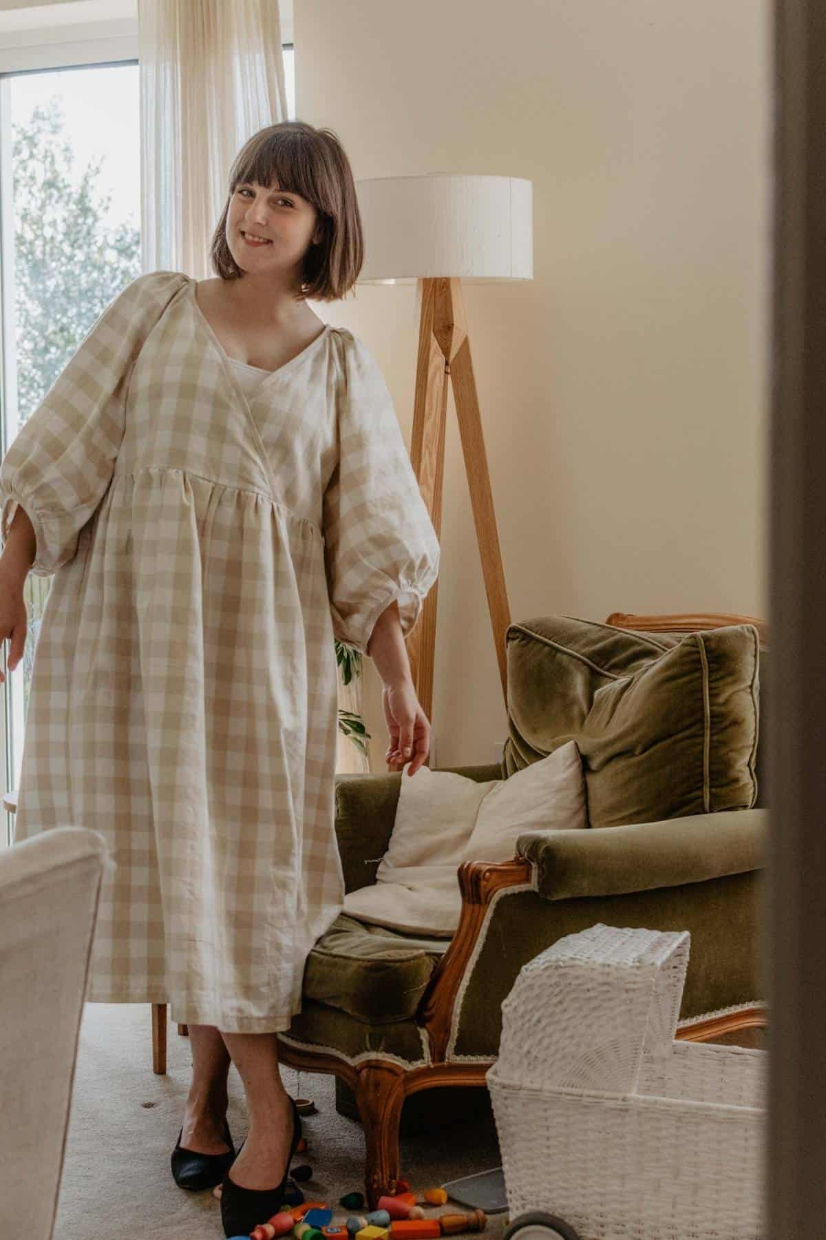 The woman in a checkered dress stands in a living room, embodying spring style with her capsule wardrobe choice. Beside her is a green upholstered chair, flanked by a floor lamp and scattered toys. She smiles warmly, creating a cozy and inviting atmosphere thats nursing and maternity friendly.