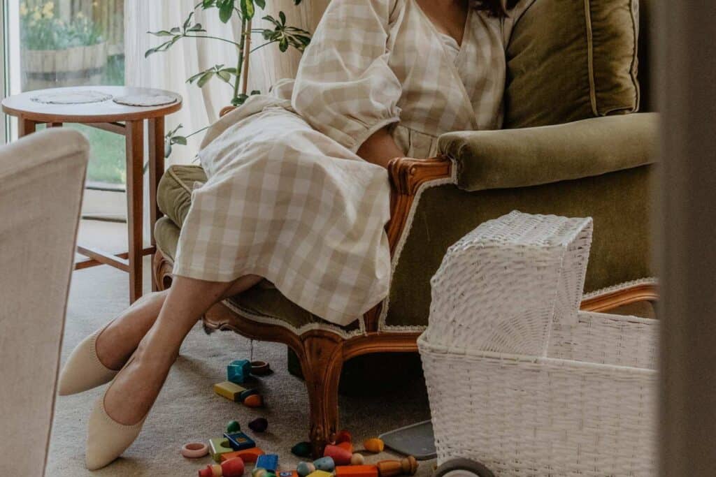 A person in a checkered dress from their capsule wardrobe sits in an armchair by a window overlooking lush spring greenery. Toys and colorful blocks are scattered on the floor beside a white wicker basket. The scene feels cozy and softly lit.