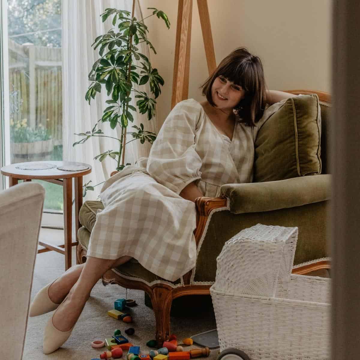 A woman with a short bob haircut lounges on a green armchair in a cozy living room. Clad in a nursing and maternity-friendly checkered dress, she relaxes amidst scattered toys. A plant basks by the large window, hinting at the freshness of spring.