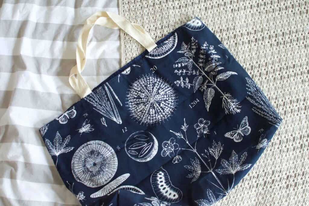 A navy blue tote bag with cream handles, showcasing white botanical and geometric designs like plants, flowers, and butterflies, rests on a beige textured surface next to a white and gray striped fabric. 