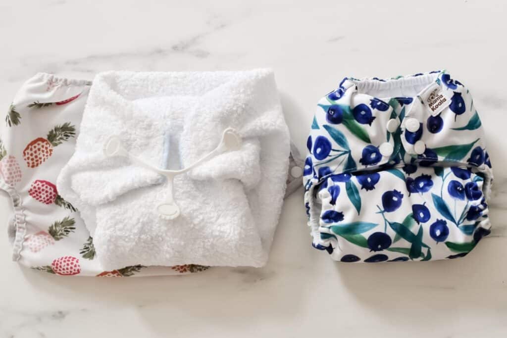 Three cloth nappies are displayed on a white surface: one with pineapple prints, a white fluffy one with a fastener, and another with blueberries print. Start using these eco-friendly alternatives for both style and sustainability in your babys daily routine.