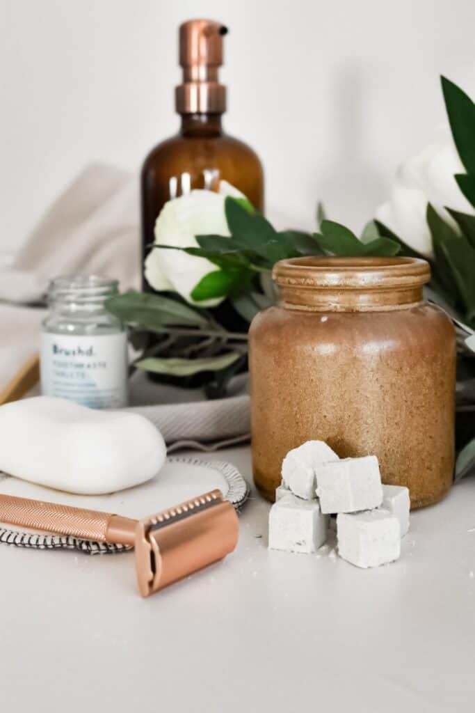 An eco-friendly bathroom setup features a copper razor, a white soap bar, and a small jar of essential dental tablets. A brown glass pump bottle completes the scene. In the foreground are white bath bombs, while green leaves and white flowers grace the background.