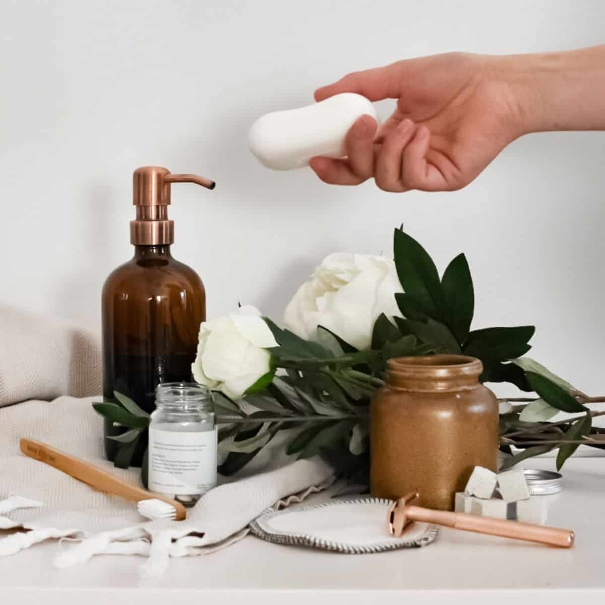 A hand presents a white soap bar over a bathroom counter adorned with eco-friendly bathroom products. The setup includes a brown soap dispenser, two white roses, a leafy branch, brown and white jars, a bronze razor, a toothbrush, and a round mirror. A light fabric is draped nearby.