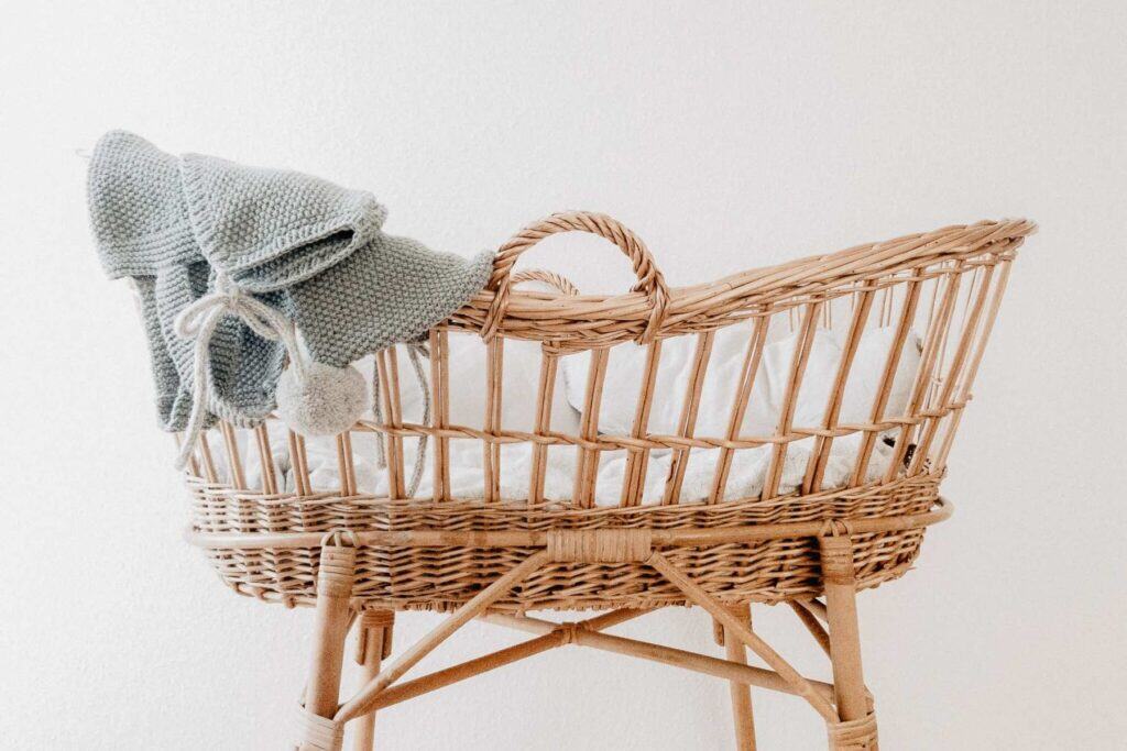 A wicker cradle with a soft white cushion rests against a plain backdrop. Draped over the side is a folded, knitted gray blanket with pom-pom ties—perfect for your little one. Opting for such pieces is one of many tips for crafting a sustainable bedroom environment.