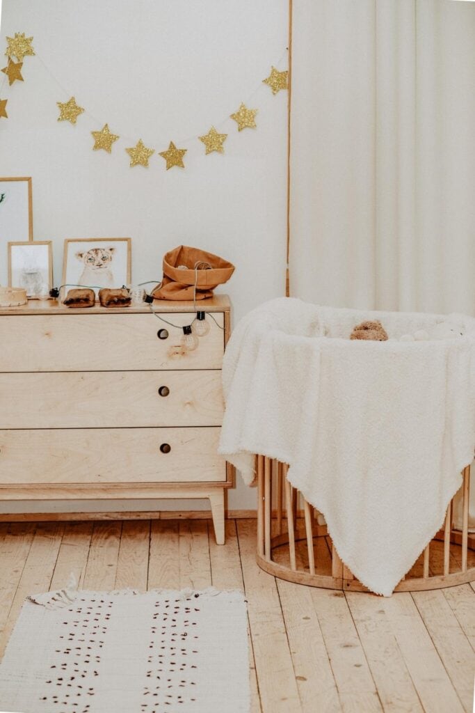 A warm-toned nursery for your little one features a wooden dresser with charming decor, framed pictures, and tiny shoes. Nearby, a cozy round bassinet holds a plush toy and fluffy blanket. Sustainable touches include gold star decorations hanging overhead.